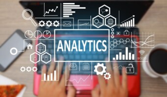 Manos escribiendo en una computadora portátil con varios íconos de análisis digital, incluidos gráficos, tablas y lupas, superpuestos a la imagen. La palabra "Analytics" se muestra de manera destacada en el centro, lo que hace alusión a las funciones avanzadas de Google Analytics 4.