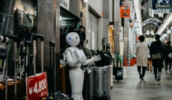 Un robot humanoide se encuentra entre el equipaje en una galería comercial con gente caminando. Un cartel de precio en primer plano dice "¥4.800". El robot, impulsado por chatbots de IA, parece listo para ayudar a los compradores con sus consultas.