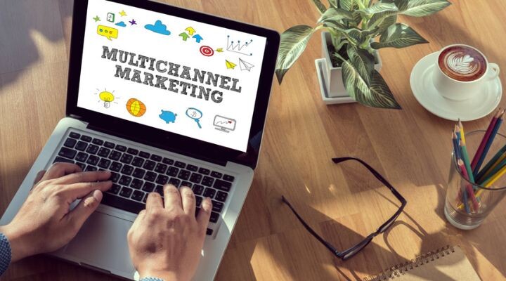 Persona escribiendo en una computadora portátil que muestra “Marketing Multicanal” en la pantalla, ilustrando estrategias de medios, con una planta, una taza de café, anteojos y lápices de colores cuidadosamente ordenados sobre un escritorio de madera.