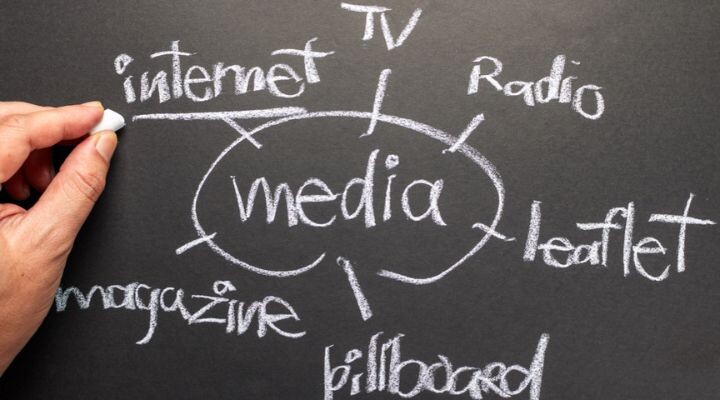 Una mano escribe “medios” en una pizarra, con ramas que se conectan a palabras como internet, televisión, radio, folleto, cartelera y revista, cada una con tiza, visualizando estrategias de medios.