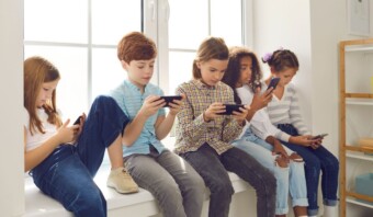 Cinco niños de generación Alfa están sentados en el alféizar de una ventana, cada uno de ellos enfocado en un teléfono inteligente. Parecen estar involucrados en actividades individuales. La habitación está bien iluminada a través de la ventana que hay detrás de ellos.