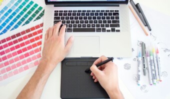 Una persona utiliza una computadora portátil y una tableta gráfica, rodeada de muestras de color, papel para dibujar, bolígrafos y marcadores, elaborando meticulosamente la identidad visual de una marca.