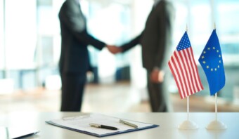 Dos personas con traje se dan la mano al fondo. En primer plano, hay una bandera de Estados Unidos, una bandera de la UE y un documento con dos bolígrafos sobre un escritorio, que simboliza un importante acuerdo político.