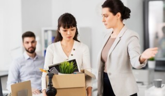 Una mujer, que lleva una caja de material de oficina, parece salir de su lugar de trabajo mientras un colega le indica la salida. Otro colega mira al fondo.