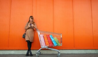 Una mujer con un vestido marrón se apoya en un carrito de compras lleno de bolsas de colores, revisando su teléfono contra un fondo de pared naranja: un momento de experiencias sin fricciones en medio de su día.