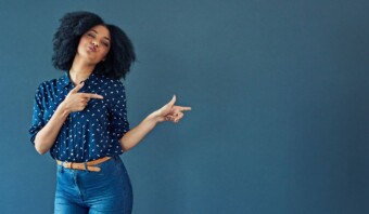 Una persona con cabello rizado que viste una camisa estampada y jeans señala con ambas manos hacia su izquierda mientras hace una expresión juguetona contra un fondo liso.