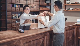 Un comerciante parado detrás de un mostrador de madera le entrega una bolsa de compras a un cliente en una tienda con estantes de cajas de zapatos y accesorios al fondo.