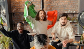 Cuatro personas celebran en el interior. Una persona ondea una bandera, otra sostiene un plato de bocadillos. El ambiente es animado, que recuerda a un evento de marketing deportivo.