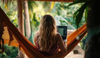 Una persona con cabello largo se sienta en una hamaca naranja con una computadora portátil en su regazo, situada al aire libre en medio de una exuberante vegetación verde, disfrutando de su zona de confort.
