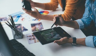 Dos personas trabajan en un proyecto en un escritorio. Una usa una tableta y un lápiz para editar una imagen, mientras que la otra sostiene una cámara. Hay fotografías y muestras de color esparcidas sobre la mesa.