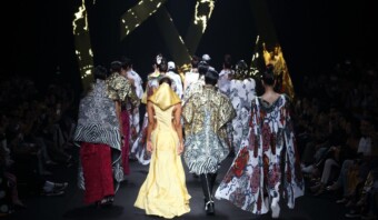 Un grupo de modelos camina por una pasarela, exhibiendo diversas prendas elaboradas y coloridas que recuerdan la extravagancia de la Gala del Met. El fondo presenta diseños abstractos iluminados.