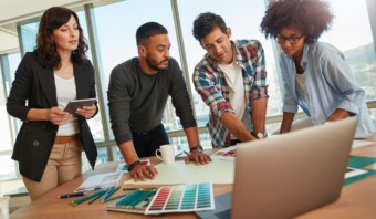 Cuatro colegas están reunidos alrededor de una mesa con computadoras portátiles, papeles y lápices de colores, colaborando en un proyecto en una oficina luminosa y moderna con grandes ventanas.