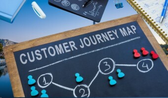 Una pizarra con la etiqueta "Mapa del recorrido del cliente" presenta un diagrama de flujo con etapas numeradas y piezas de juego rojas y azules que representan las posiciones de los clientes. Cerca hay papeles, un bolígrafo y una lupa.