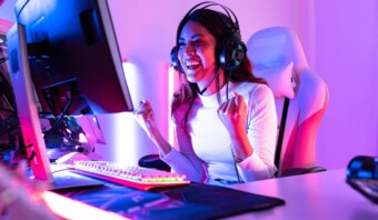 Un jugador de pelo largo con auriculares está sentado en una silla de juego, mirando la pantalla de una computadora, apretando los puños y sonriendo. La habitación está iluminada con luces rosas y azules.