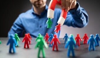 Una persona con una camisa azul usa un imán rojo y blanco para recoger una pequeña figura verde de un grupo de figuras rojas, verdes y azules sobre una mesa, de forma muy similar a la orientación precisa que se ve en la generación de leads.
