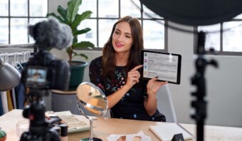 Una microinfluencer se sienta en un escritorio con artículos de maquillaje, apuntando a la pantalla de una tableta mientras graba un video con una cámara.