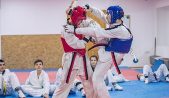 Dos artistas marciales vestidos con equipo de protección participan en un combate de entrenamiento mientras los espectadores sentados observan en un dojo, lo que recuerda la disciplina y la emoción que anticipamos en París 2024.