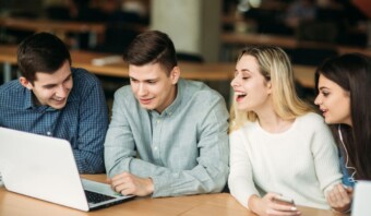 Cuatro adultos jóvenes están reunidos alrededor de una computadora portátil y participan en una conversación. Están sentados en una mesa de madera en una habitación con otras mesas y sillas.