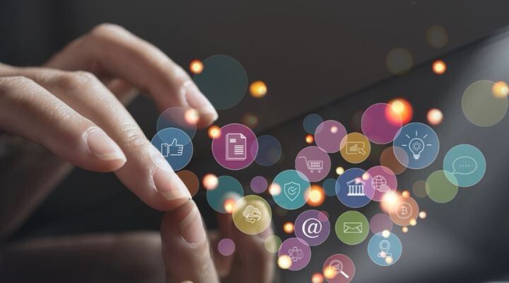 Primer plano de manos interactuando con una pantalla táctil, con varios íconos coloridos como pulgares hacia arriba, sobre y engranaje flotando alrededor, representando la comunicación digital y la tecnología mientras se optimiza el presupuesto.