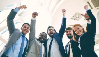 Los colaboradores, vestidos con ropa de negocios, levantan alegremente sus brazos en señal de celebración, de pie en el interior bajo un techo brillante.