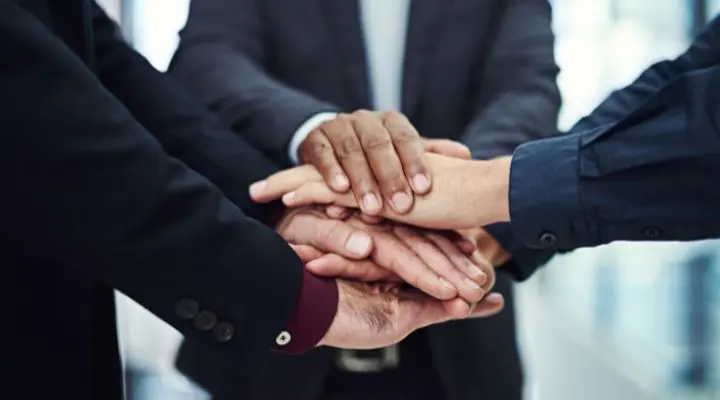 Varias manos apiladas una sobre otra, simbolizando el trabajo en equipo y la colaboraciÃ³n entre los colaboradores. Las personas visten ropa formal de negocios.