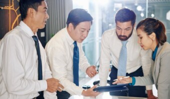 Cuatro profesionales de negocios con camisa blanca y corbata están reunidos alrededor de una mesa, enfrascados en una discusión seria. Uno de ellos, centrado en la gestión de crisis, sostiene una tableta mientras otro la señala.