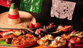 Una mesa está preparada con varios platos mexicanos, un sombrero y decoraciones tradicionales que incluyen papel picado y una manta colorida.