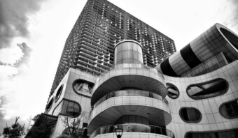 Una fotografía monocromática de un edificio moderno de varios pisos con formas circulares y rectangulares que sobresalen, con amplios paneles de vidrio y un patrón similar a una cuadrícula en los niveles superiores, encarna un diseño arquitectónico elegante adecuado para campañas de marketing innovadoras.