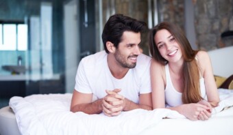 Un hombre y una mujer, ambos vestidos con camisas blancas, yacen en una cama, sonriendo y mirándose con sexualidad positiva en un dormitorio bien iluminado.