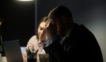 Dos personas están sentadas en una habitación poco iluminada y parecen estresadas. Una se cubre la cara con una mano y la otra parece consolarlas. Sobre el escritorio hay una computadora portátil y una taza.