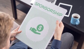 Una persona sostiene un folleto de patrocinio que presenta un símbolo de dinero vibrante en la portada, junto a una taza de café humeante sobre la mesa: se trata de inversión publicitaria.