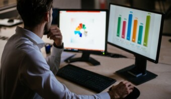 Una persona se sienta en un escritorio y analiza gráficos de datos en dos monitores de computadora. En uno de ellos se muestra un gráfico circular colorido y en el otro, gráficos de barras.