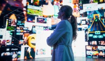 Una mujer se encuentra de pie, segura, frente a una pantalla grande que muestra varias imágenes y vídeos digitales, encarnando la esencia de su estrategia de medios.