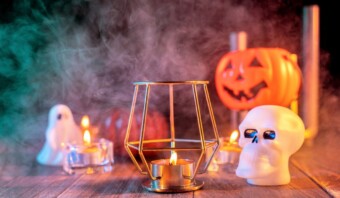 Una vela encendida parpadea en un soporte de metal, envuelta por decoraciones de Halloween como una calavera, una calabaza y un fantasma, con humo místico arremolinándose en el fondo.