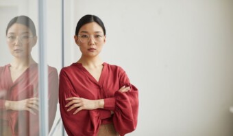Una mujer con blusa roja y gafas aparece de pie con los brazos cruzados, encarnando la esencia de la industria creativa, mientras su reflejo brilla en la superficie del vidrio.