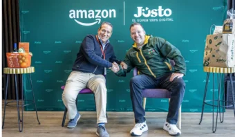Dos hombres se dan la mano sentados en sillas frente a un telón de fondo con la marca Amazon y Jüsto. Bolsas de la compra y una caja adornan las mesitas que hay junto a ellos.
