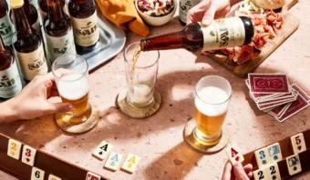 Se ven manos jugando con fichas y cartas sobre una mesa. Aunque es el Día Mundial sin Alcohol, se sirven cervezas en vasos y hay botellas y bocadillos cerca.