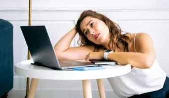 Una mujer apoya la cabeza en su mano en una mesa redonda, mirando la pantalla de una computadora portátil con una expresión pensativa.