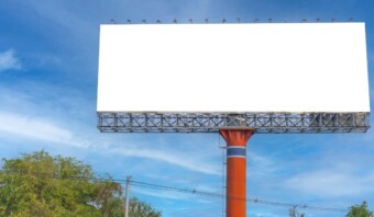 Un gran cartel vacío sobre un poste rojo alto se alza contra el cielo azul claro, rodeado de unos pocos árboles. Evoca una sensación de "mundo sin publicidad", que invita a la contemplación de nuestro paisaje visual sin publicidad.