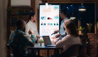 Cuatro personas en una oficina con poca luz analizan datos de personas mientras discuten sobre ellos en una gran pantalla digital. Una persona señala la pantalla, enfatizando los datos, mientras que otra toma notas detalladas en una computadora portátil.
