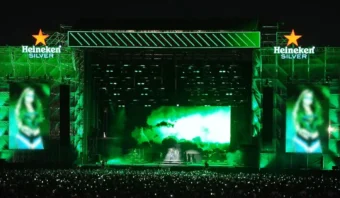 Un gran escenario de conciertos, iluminado en verde, muestra la marca Heineken Silver en una pantalla gigante, con una multitud de personas capturando el momento con entusiasmo (no es necesario The Boring Phone aquí).