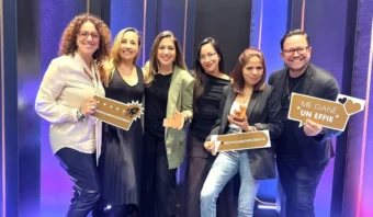 Un grupo de seis personas se encuentran juntas, sosteniendo premios y carteles, sonriendo a la cámara. El fondo presenta luces azules verticales que celebran el éxito de VML México en Effie 2024.
