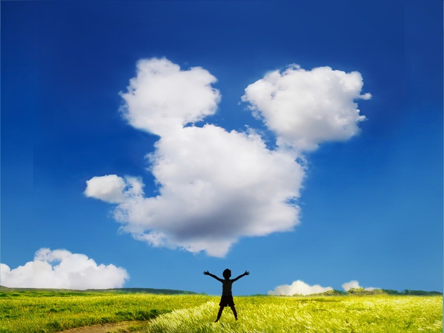 Una persona está parada en un campo de hierba con los brazos levantados, mirando una formación de nubes que se asemeja a la silueta de un ratón en un cielo azul claro.