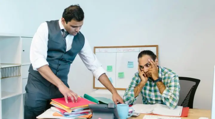 Dos hombres en una oficina: uno señala unos archivos sobre un escritorio y el otro está sentado con un teléfono y apoya la cabeza en la mano. Al fondo hay una pizarra con notas adhesivas Síndrome del boreout laboral