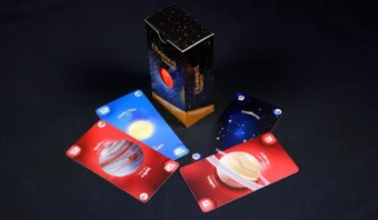Un juego de cartas con temática espacial, que presenta innovación en impresión con IA, se extiende con una caja de cartas abierta en el centro sobre una superficie negra.