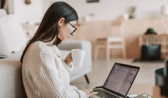 Una persona sentada en el suelo, usando una computadora portátil y sosteniendo una taza en una habitación acogedora.