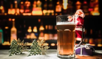 Una jarra de cerveza en el mostrador del bar con una pequeña figura de Papá Noel, árboles decorativos y una caja de regalo cerca.