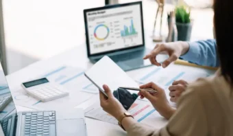 Dos personas están enfrascadas en una discusión sobre informes comerciales en un escritorio, una de ellas sostiene una tableta y señala con un bolígrafo, mientras que en la pantalla de una computadora portátil se ven gráficos que brillan en el fondo. Su objetivo es crear contenido imperecedero que siga siendo valioso y relevante a lo largo del tiempo.