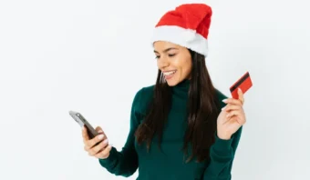 Una mujer con un gorro de Papá Noel sonríe mientras sostiene un teléfono inteligente y una tarjeta de crédito digital sobre un fondo blanco.