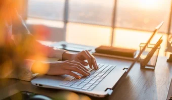 Persona escribiendo en una computadora portátil en un escritorio, con una vista del atardecer a través de grandes ventanas en el fondo.